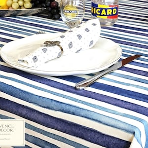 May include: A white dinner plate sits on a blue and white striped tablecloth. A white napkin with a small blue geometric pattern is held by a metal napkin ring. A silver knife rests on the tablecloth next to the plate. A glass with the text RICARD is visible.