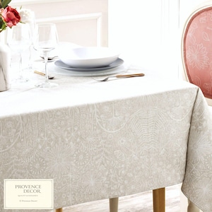 May include: A beige tablecloth with a detailed white floral pattern covers a wooden table. White plates, bowls, and wine glasses are set on the table. The text "PROVENCE DECOR QUALITY & ELEGANCE" is in the lower left corner.
