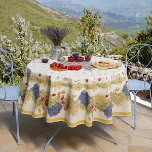 May include: A white tablecloth with a floral pattern featuring lavender, wheat, and berries. The tablecloth is laid on a round table with two blue metal chairs. The table is set with a bowl of fruit, a plate of cookies, and a vase of lavender.