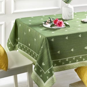 May include: A green tablecloth with a white heart pattern and a decorative border. The tablecloth is draped over a table, with a white plate of pink flowers and a white vase with flowers on top.