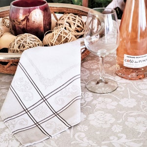 May include: A white linen napkin with a brown border and a floral pattern is folded on a beige tablecloth with a floral pattern. A glass of wine, a bottle of wine labelled "Gerard Bertrand Cote des Roses", and a wicker basket with decorative balls are on the table.