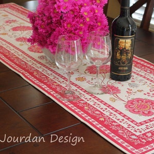 May include: A white table runner with a pink floral pattern and red borders. Two wine glasses and a bottle of wine are on the runner, which is on a dark wood table.
