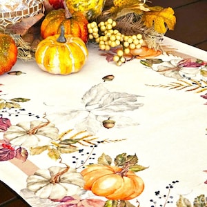 May include: A cream-colored table runner featuring a printed pattern of orange and white pumpkins, brown acorns, and autumn leaves. The runner is decorated with a border of botanical illustrations and is topped with several small decorative pumpkins.