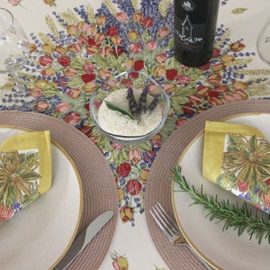 May include: A table setting with two place settings, each with a white plate, a yellow napkin with a floral design, a fork, a knife, and a sprig of rosemary. The table is covered in a white tablecloth with a floral pattern. There is a small bowl of rice and lavender in the center of the table.