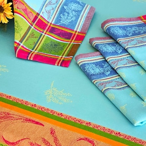 May include: A collection of colorful, patterned kitchen linens. The image features a checkered napkin with green, pink, and yellow squares, and several folded blue napkins with floral designs. The linens are arranged on a light blue surface.