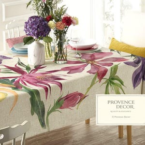 May include: A beige tablecloth with a large floral design in shades of pink, purple, and yellow. The table is set with plates, glasses, and vases of flowers. The tablecloth has the text "PROVENCE DECOR" in a decorative frame.