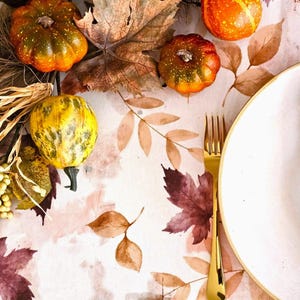 May include: A seasonal table setting featuring small pumpkins in orange, yellow, and green. The table is decorated with autumn leaves and a gold fork next to a white plate. The tablecloth has a watercolour leaf pattern.