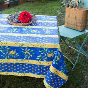 May include: A blue and yellow striped tablecloth with a pattern of olive branches and small yellow flowers. The tablecloth is laid out on a table outdoors with a red flower arrangement in the center.