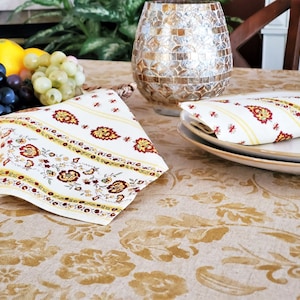 May include: A white linen tablecloth with a gold floral pattern. Two napkins with a yellow and red floral pattern are folded on the table. One napkin is on a plate, and the other is folded on the table.