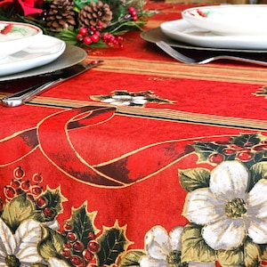 May include: A festive red tablecloth with a floral and ribbon design, set for a holiday meal. White plates, silver chargers, and silverware are arranged on the table, with pinecones and greenery in the background.