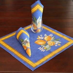 Puede incluir: Un mantel individual y servilletas azules con un dise&ntilde;o de lim&oacute;n. El mantel individual cuadrado tiene un borde amarillo. Las servilletas est&aacute;n dobladas y colocadas sobre el mantel individual. Un vaso transparente est&aacute; en el centro. El juego de mesa est&aacute; sobre una mesa de madera marr&oacute;n oscuro.