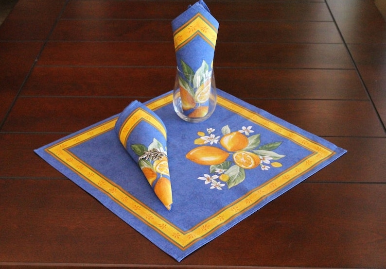 Puede incluir: Un mantel individual y servilletas azules con un dise&ntilde;o de lim&oacute;n. El mantel individual cuadrado tiene un borde amarillo. Las servilletas est&aacute;n dobladas y colocadas sobre el mantel individual. Un vaso transparente est&aacute; en el centro. El juego de mesa est&aacute; sobre una mesa de madera marr&oacute;n oscuro.