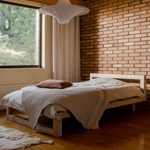 Peut inclure: Un lit en bois minimaliste avec une couverture et un oreiller clairs. Une couverture texturée de couleur claire est drapée sur le lit. La pièce comprend un mur de briques et une grande fenêtre avec vue sur des arbres. Le lit mesure environ 200 cm de long.