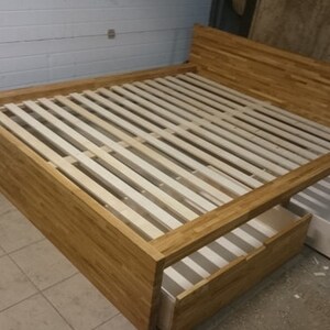Peut inclure: Un cadre de lit en bois avec une tête de lit et un pied de lit à lattes. Le lit a deux tiroirs qui sont partiellement ouverts, révélant un intérieur blanc.