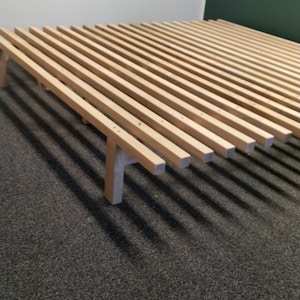 Puede incluir: Un somier de cama de listones de madera con un acabado natural. El marco está hecho de múltiples listones que están conectados por una serie de soportes de madera. El marco está sentado en una alfombra gris oscuro.