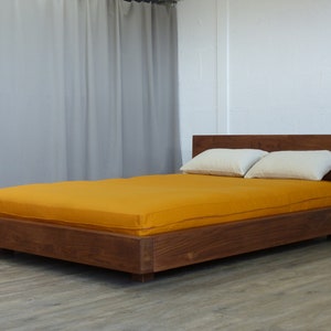 Op de afbeelding: Een houten platform bedframe met een gele matras en twee witte kussens. Het bedframe is gemaakt van donker hout en heeft een eenvoudig, modern ontwerp.