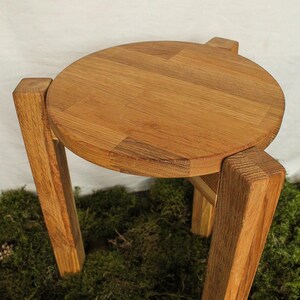 Puede incluir: Un taburete redondo de madera con cuatro patas. El taburete está hecho de madera marrón claro y tiene un acabado liso y pulido.