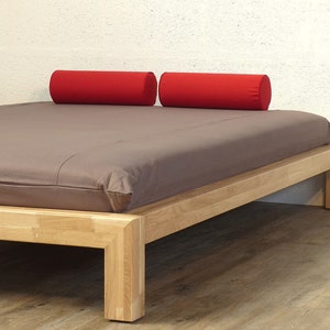 Könnte beinhalten: Ein Holzbettgestell mit einer grauen Matratze und zwei roten zylindrischen Kissen. Das Bettgestell hat ein schlichtes, modernes Design mit einem niedrigen Profil.