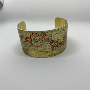 May include: Wide, open cuff bracelet with a cream-colored base. The bracelet features a colorful, abstract design with red, yellow, blue, and green swirls. The design is reminiscent of a watercolor painting. The bracelet is made of a metal material.