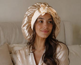 May include: A champagne-colored satin pajama set consisting of a long-sleeved button-down shirt and matching trousers, paired with a matching pleated satin sleep bonnet featuring a decorative bow detail on the front and an elasticized band.