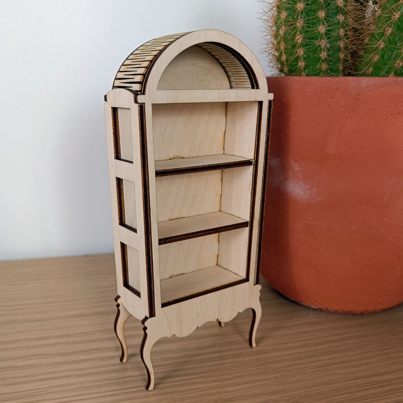 May include: A wooden display cabinet with an arched top and three shelves. The cabinet has a light wood finish and dark brown trim. It has decorative legs and is standing on a wooden surface.