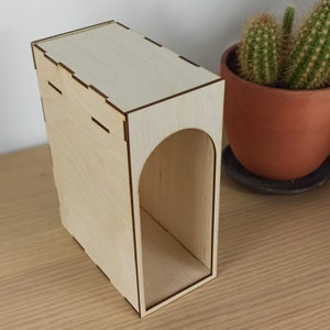 May include: A wooden box with a cutout archway. The box is made of light wood and has a natural finish. The archway is cut out of the front of the box and is shaped like a rounded rectangle.