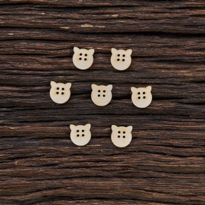 Peut inclure: Sept boutons en bois en forme de tête de cochon avec deux petits trous pour coudre. Les boutons sont disposés de manière symétrique sur une surface en bois brun foncé.