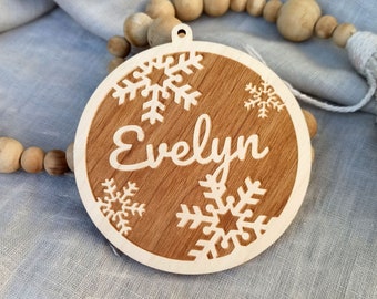 May include: A round wooden bauble with a brown stain and a white snowflake design. The bauble has the name "Evelyn" engraved on it.