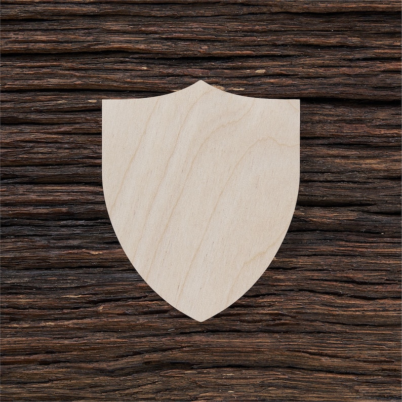 May include: A plain wooden shield shape, cut out from a piece of wood. The shield is light brown and has a smooth surface. It is sitting on a dark brown wooden background.