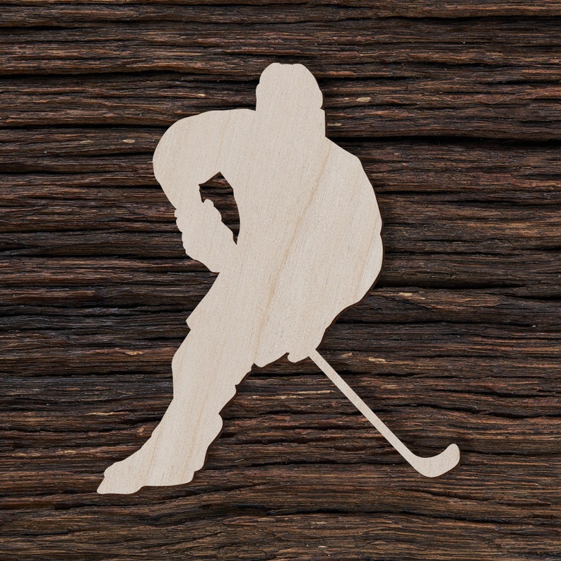 May include: A wooden cutout of a hockey player in a skating pose, holding a hockey stick. The cutout is light-colored and set against a dark brown, textured wooden background. The player is in motion, ready to shoot the puck.