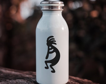 May include: A white water bottle with a silver cap. A black Kokopelli figure is printed on the front. The bottle is cylindrical and appears to be made of metal. The background is blurred, with a natural, outdoor setting.