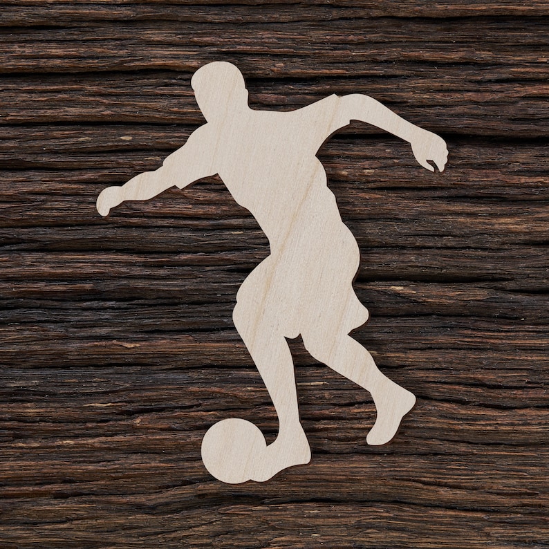 May include: A white wooden silhouette of a soccer player kicking a ball. The silhouette is cut out and placed on a dark brown wooden background.