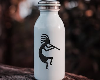 May include: A white insulated water bottle with a silver lid. The bottle features a black Kokopelli design. The bottle is cylindrical and appears to be made of stainless steel. The background is blurred, with natural tones of brown and green.