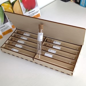 May include: A wooden box with a hinged lid, containing multiple glass tubes with cork stoppers. Each tube has a white label with text and a sprig design. One tube stands upright, while others are arranged horizontally. The box is open, revealing the contents.