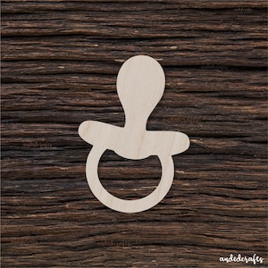 Puede incluir: Un recorte de madera de un chupete, un juguete para bebés, con forma de chupete tradicional con un anillo redondo y una tetina. El recorte es blanco y está sobre un fondo de madera marrón.