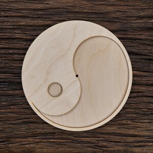 Könnte beinhalten: Ein hölzernes Yin-Yang-Symbol mit einem kleinen Loch in der Mitte. Das Symbol ist aus hellbraunem Holz gefertigt und befindet sich auf einem dunkelbraunen Holzgrund.