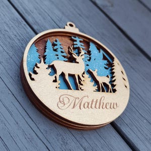 Pode incluir: Enfeite de Natal de madeira com a silhueta de um veado e um veado num cenário de floresta. O enfeite redondo apresenta um design em camadas com tons azuis e castanhos. O nome "Matthew" está gravado na parte inferior.