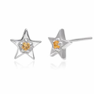May include: A pair of star-shaped stud earrings crafted from a polished silver-toned metal. Each earring features a central, round, faceted yellow gemstone set within a prong setting at the centre of the star, complete with a standard post backing.