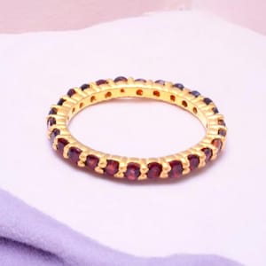 以下が含まれることがあります： 小さな丸いダークレッドの宝石が連続して並んだ、金色のリング。 白と薄紫色の背景に配置され、リングのデザインを際立たせています。