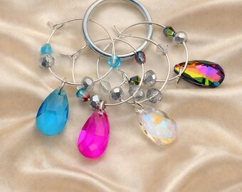 May include: A set of silver-toned metal wine glass charms featuring teardrop-shaped crystal pendants in blue, pink, iridescent white, and multicolored hues. Each charm is accented with small round beads and attached to a circular silver ring.