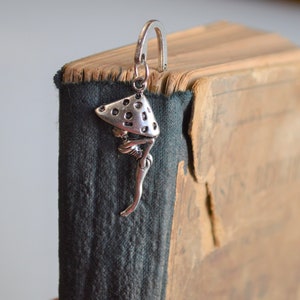 May include: Silver bookmark featuring a stylised figure with a mushroom-shaped hat. The bookmark is attached to an old, worn book.