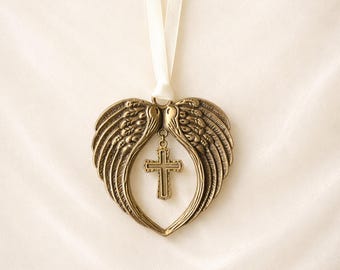 May include: A bronze-toned metal ornament shaped like a heart formed by two feathered angel wings. A small, matching bronze-toned cross hangs from the center of the wings. The entire piece is suspended by a thin, cream-colored satin ribbon.