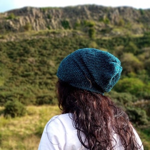 May include: A teal knitted beanie hat. The hat is worn by a person with long, dark hair and a white shirt. The background features a blurred landscape of green hills and a rocky cliff.