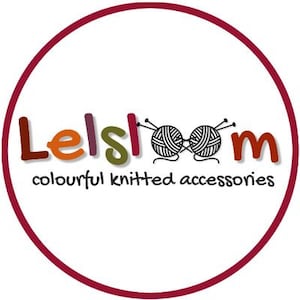 以下が含まれることがあります： マルーンの縁取りのある円形のロゴ。カラフルな文字で「Lelsloom」の文字と、毛糸玉と針のペアが描かれています。下には「colourful knitted accessories」と書かれています。