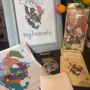 May include: A framed cross-stitch map of North America with the text 'my travels' in a light blue frame. Beside it is a North America cross-stitch map kit in a plastic sleeve, a printed pattern sheet, a bundle of colorful embroidery thread, and fabric.