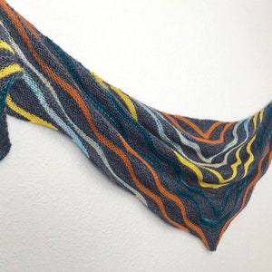 Könnte beinhalten: Ein grauer Strickschal mit wellenförmigen Streifen in Gelb, Orange, Türkis und Hellblau. Der Schal ist dreieckig und scheint handgefertigt zu sein. Die Farbpalette ist lebendig und das Design ist auffällig.
