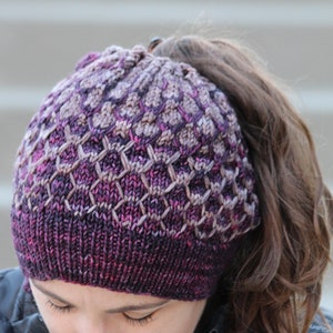 Può includere: Un cappello di lana viola e marrone con un foro per la coda di cavallo. Il cappello ha un motivo testurizzato ed è fatto con un filato morbido.