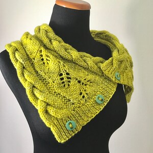 Op de afbeelding: Een groene gebreide cowl met een kabelbreipatroon en twee groene knopen. De cowl wordt getoond op een zwart manikin.
