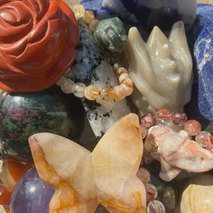 May include: A collection of various gemstone carvings and beaded bracelets. The carvings include a red rose, a butterfly, and a stylized flower. The bracelets feature orange and clear beads. Other stones include green, white, and blue.