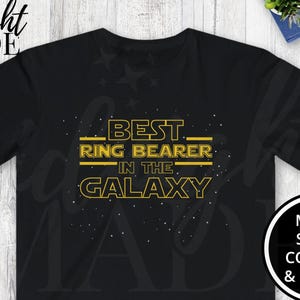 May include: Black t-shirt with the text "BEST RING BEARER IN THE GALAXY" in yellow. The shirt is displayed on a light wood-grain surface, with a pair of blue and gray sneakers and a small potted plant visible.